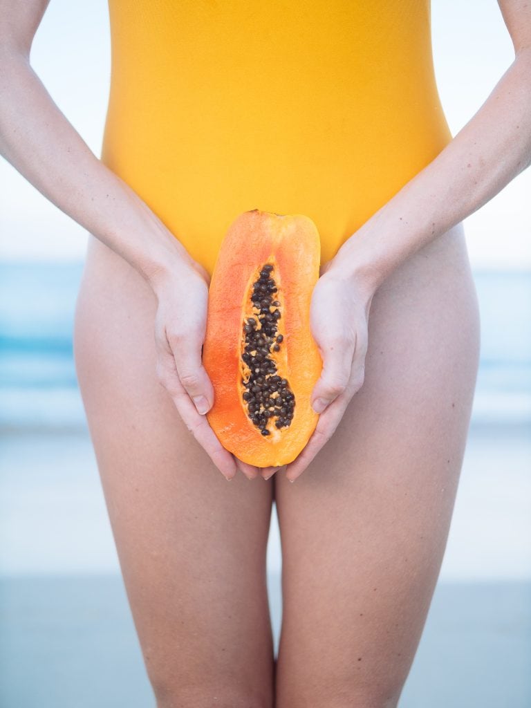 A person in a yellow swimsuit holds half a papaya in front of their waist, with a beach in the background.