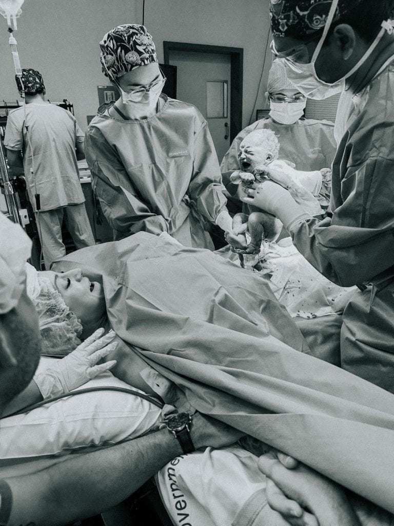 Medical professionals in a delivery room assist in the birth of a baby. The newborn is held up by one of the medical staff, while the mother lies in bed covered by a surgical drape.