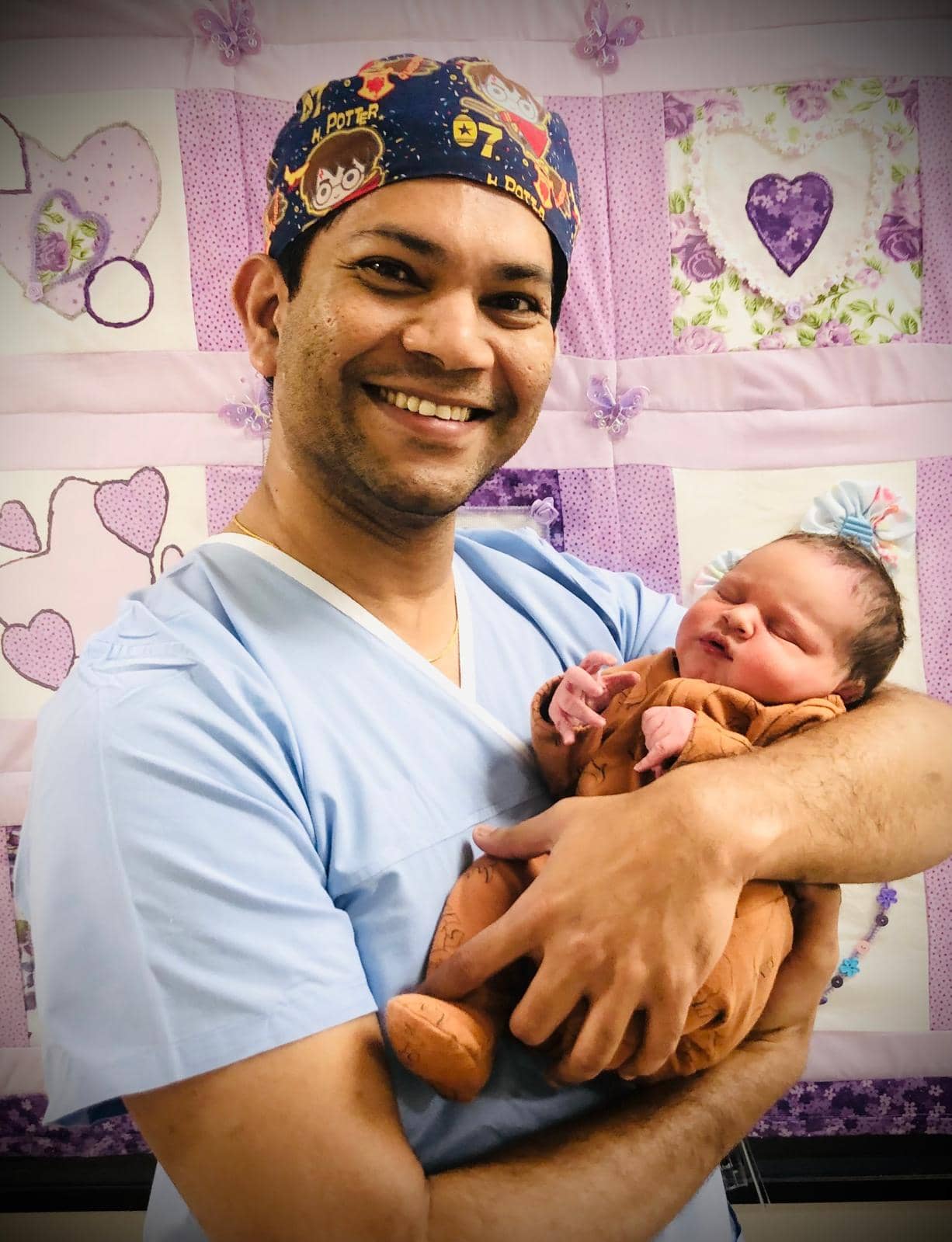 A person wearing medical scrubs and a colorful surgical cap is smiling and holding a newborn baby wrapped in a brown blanket in front of a quilted purple and pink backdrop.